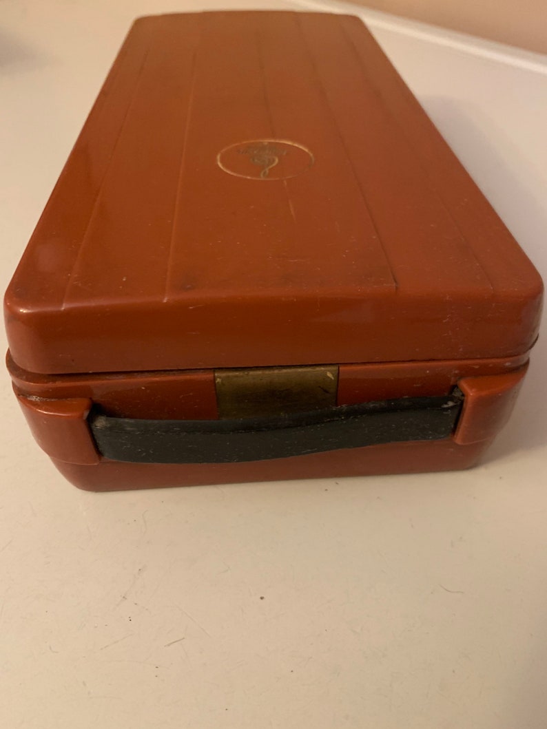 Puede incluir: Un estuche rectangular marr&oacute;n con asa negra y cierre de lat&oacute;n. La caja tiene una textura acanalada en la parte superior y un peque&ntilde;o emblema ovalado. La caja est&aacute; cerrada y parece estar hecha de pl&aacute;stico duro o material compuesto.