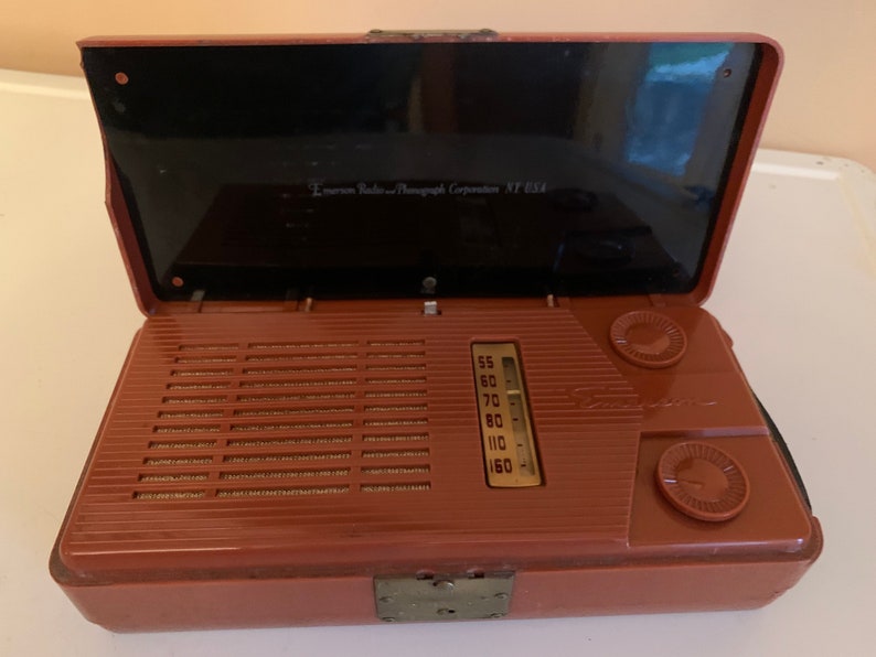 Puede incluir: Radio Emerson vintage en una caja marr&oacute;n con tapa negra. La radio tiene una rejilla de altavoz, dial de sintonizaci&oacute;n y dos perillas de control. El texto "Emerson Radio and Phonograph Corporation NY USA" es visible.