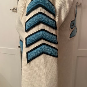 May include: White knitted sweater with blue and black chevron patches on the sleeve.