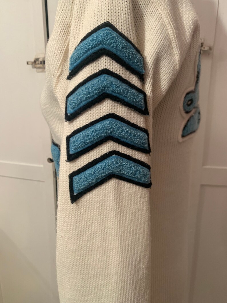 May include: White knitted sweater with blue and black chevron patches on the sleeve.