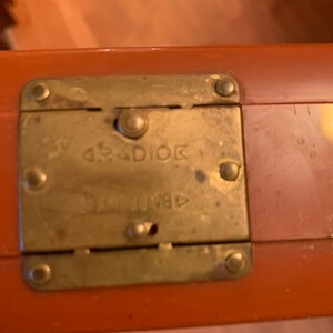 Puede incluir: Primer plano de un cierre de maleta naranja vintage. El cierre de lat&oacute;n es rectangular con esquinas redondeadas y est&aacute; asegurado con cuatro remaches. La palabra "RADIOK" est&aacute; estampada en el cierre.