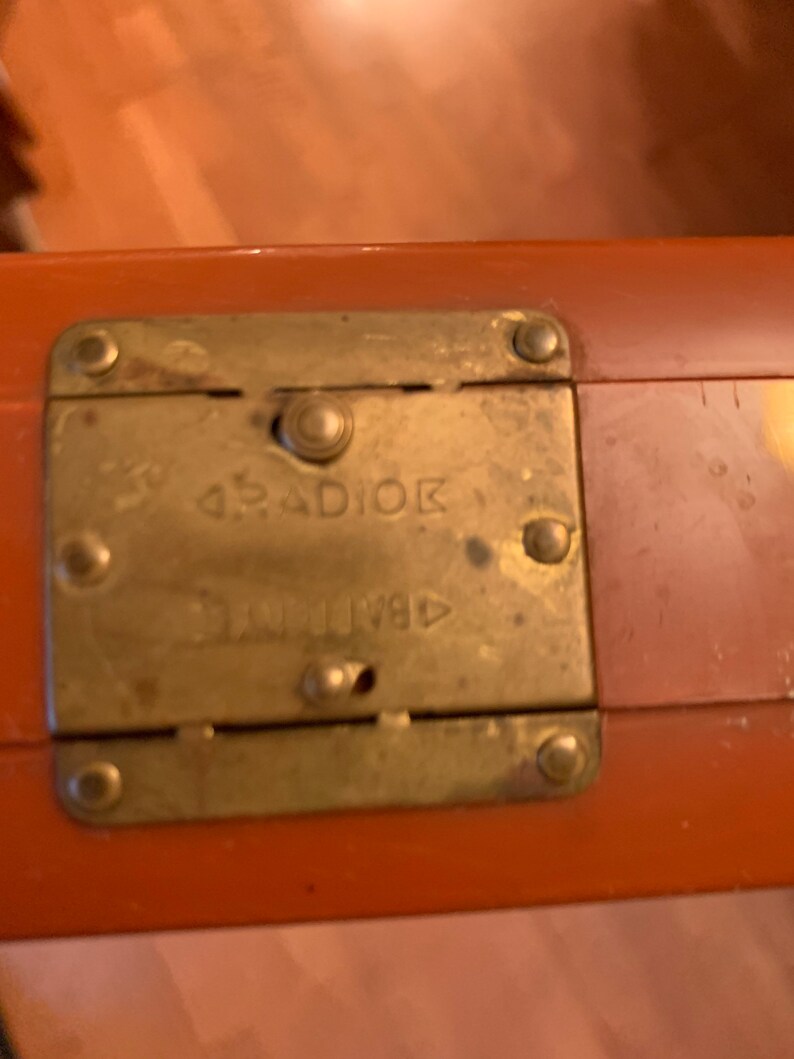 Puede incluir: Primer plano de un cierre de maleta naranja vintage. El cierre de lat&oacute;n es rectangular con esquinas redondeadas y est&aacute; asegurado con cuatro remaches. La palabra "RADIOK" est&aacute; estampada en el cierre.