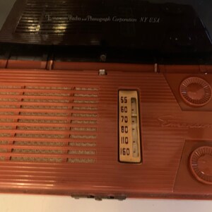 Puede incluir: Radio Emerson vintage en una carcasa marr&oacute;n rojiza, abierta para revelar el dial y el altavoz. La radio tiene forma rectangular con una rejilla de altavoz y dos perillas circulares. El dial muestra los n&uacute;meros de frecuencia de radio. El logotipo de Emerson es visible.