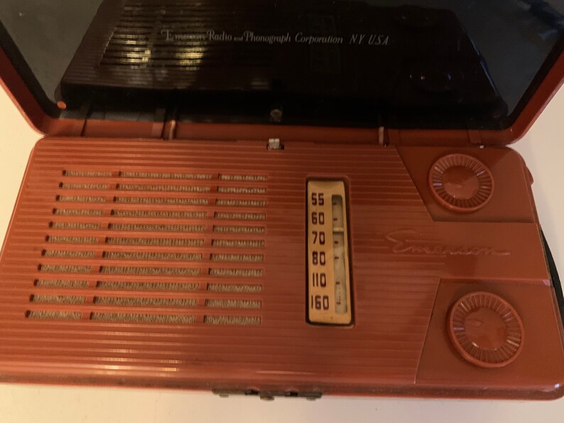 Puede incluir: Radio Emerson vintage en una carcasa marr&oacute;n rojiza, abierta para revelar el dial y el altavoz. La radio tiene forma rectangular con una rejilla de altavoz y dos perillas circulares. El dial muestra los n&uacute;meros de frecuencia de radio. El logotipo de Emerson es visible.