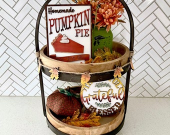Könnte beinhalten: Ein zweistufiges dekoratives Tablett mit herbstlichen Dekorationen. Die obere Ebene zeigt ein Schild mit der Aufschrift "Homemade Pumpkin Pie" und einen kleinen Strauß orangefarbener Blumen in einem grünen Glas. Die untere Ebene hat einen Kürbis und ein Schild mit der Aufschrift "Grateful Blessed".