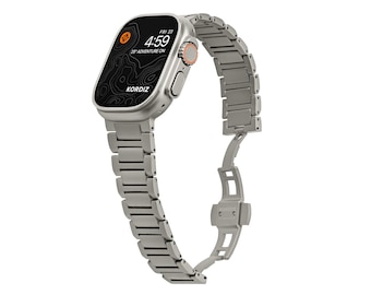May include: A silver metal Apple Watch band with a silver metal case. The watch face is black with a topographic map design and the time is 4:59. The watch band has a clasp closure.