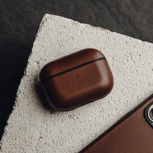 May include: A brown leather case for wireless earbuds. The case has a black seam around the edge and a subtle texture.