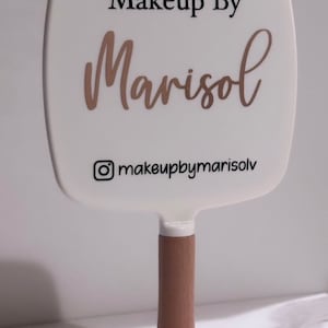 May include: White handheld makeup mirror with a wooden handle. The mirror has the text "Makeup By Marisol" and the Instagram handle "@makeupbymarisolv" printed on it.