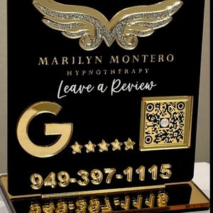 May include: A black and gold sign with the name "Marilyn Montero" and the words "Hypnotherapy" and "Leave a Review". The sign has a gold Google logo, five gold stars, a QR code, and a phone number. A gold wing design is at the top.
