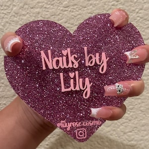 May include: Pink glitter heart-shaped sign with the text "Nails by Lily" in pink lettering. A hand is holding the sign, displaying pink and white nail tips with rhinestones.