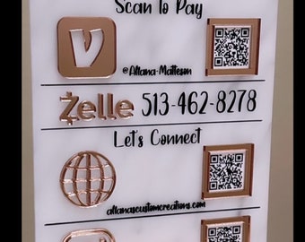 May include: A white sign with a rose gold stand, featuring a rainbow graphic, the text "Allana's Custom Creations", "Scan to Pay", "@Allana-Matteson", "Zelle 513-462-8278", "Let's Connect", "allanascustomcreations.com", and various social media icons with QR codes.