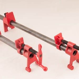 Puede incluir: Dos abrazaderas de tubo rojas con mangos ajustables. Las abrazaderas son de metal y tienen un acabado negro. Las abrazaderas se utilizan para sujetar objetos juntos.