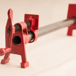 May include: A red metal pipe clamp with a black handle and a threaded screw. The clamp is open and ready to be used.