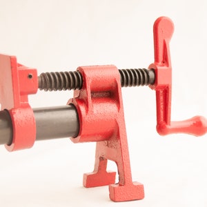 May include: A red metal pipe clamp with a black threaded screw and a lever handle. The clamp is designed to hold pipes or other objects securely in place.