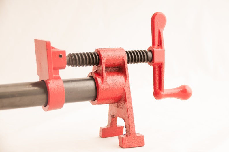 May include: A red metal pipe clamp with a black threaded screw and a lever handle. The clamp is designed to hold pipes or other objects securely in place.