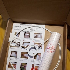 May include: A white lampshade making kit with instructions, a white metal lampshade frame, and a roll of clear adhesive-backed grid paper. The instructions include steps for attaching the fabric to the frame and trimming the excess.