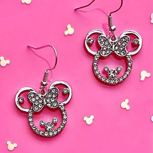 May include: A pair of silver earrings with a rhinestone-encrusted Minnie Mouse design. The earrings feature a bow and Mickey Mouse ears.