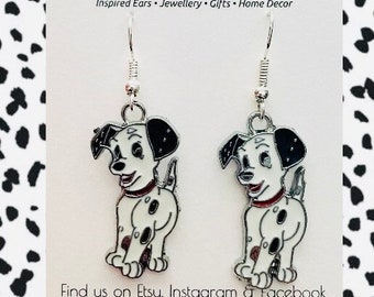 May include: A pair of silver dangle earrings featuring a cartoon Dalmatian puppy design. The earrings are white with black spots and have a red collar. The earrings are on a white background with black polka dots.