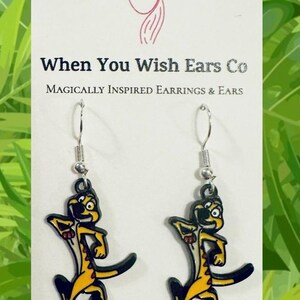 May include: A pair of dangle earrings featuring a cartoon character in yellow and black. The earrings are attached to silver-colored hooks. The card behind the earrings reads "When You Wish Ears Co" and "Magically Inspired Earrings & Ears."