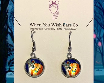 May include: A pair of silver dangle earrings with a blue background and a cartoon image of Lady and the Tramp. The earrings are on a white card with the text "When You Wish Ears Co. Inspired Ears Jewellery Gifts Home Decor. Find us on Etsy, Instagram & Facebook."