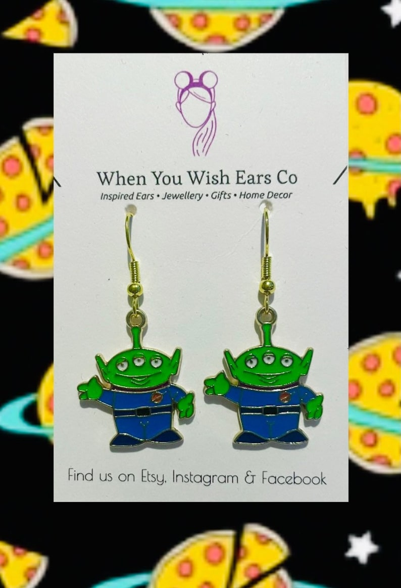 May include: A pair of gold dangle earrings featuring a green alien character with three eyes, a blue jumpsuit, and a friendly expression. The earrings are on a white card with the text "When You Wish Ears Co. Inspired Ears. Jewellery. Gifts. Home Decor. Find us on Etsy, Instagram & Facebook."