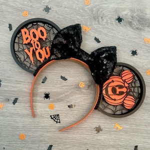 Könnte beinhalten: Halloween-Stirnband mit schwarzer Pailletten-Schleife und orangefarbenen Akzenten. Das Stirnband hat zwei kreisförmige Ohrendesigns, eines mit dem Text "Boo to You" und das andere mit einem Kürbis-Mickey-Mouse-Design. Dekorative Spinnen und Fledermäuse sind verstreut.