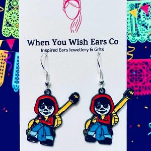 May include: A pair of dangle earrings featuring a cartoon character from the movie Coco. The character is a skeleton wearing a red shirt, blue jeans, and a yellow guitar. The earrings are silver and have a hook closure. The text "When You Wish Ears Co" and "Inspired Ears Jewellery & Gifts" is printed on the white card behind the earrings. The text "On Etsy Instagram Facebook" is printed at the bottom of the card.