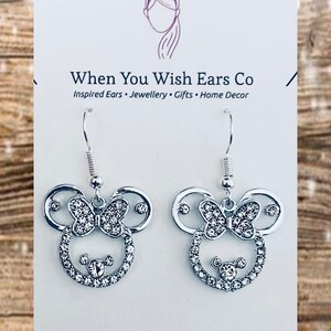 May include: Silver dangle earrings with a rhinestone-encrusted Mickey Mouse head design. The earrings feature a bow with rhinestones and a small Mickey Mouse head in the center. The earrings are on a white card with the text "When You Wish Ears Co. Inspired Ears. Jewellery. Gifts. Home Decor. Find us on Etsy, Instagram & Facebook."