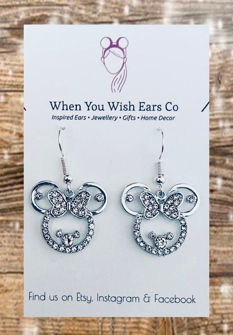 May include: Silver dangle earrings with a rhinestone-encrusted Mickey Mouse head design. The earrings feature a bow with rhinestones and a small Mickey Mouse head in the center. The earrings are on a white card with the text "When You Wish Ears Co. Inspired Ears. Jewellery. Gifts. Home Decor. Find us on Etsy, Instagram & Facebook."