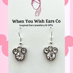 Puede incluir: Pendientes de plata con un dise&ntilde;o de cabeza de Mickey Mouse de cristal. Los pendientes est&aacute;n en una tarjeta blanca con el texto "When You Wish Ears Co. Inspired Ears Jewellery & Gifts. On Etsy Instagram Facebook."