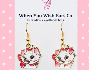 May include: A pair of gold-toned hook earrings featuring a white cat character with pink bows and accents. The earrings are displayed on a white card with the text When You Wish Ears Co Inspired Ears Jewellery & Gifts and On Etsy Instagram Facebook.