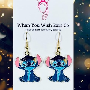 May include: Gold dangle earrings featuring a blue Stitch character wearing a graduation cap. The character has pink ears and a blue body. The earrings are on a white background with the text "When You Wish Ears Co Inspired Ears Jewellery & Gifts On Etsy Instagram Facebook".