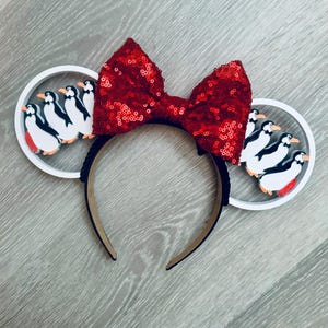 Puede incluir: Una diadema con un gran lazo de lentejuelas rojo brillante centrado entre dos marcos circulares blancos. Cada marco contiene una serie de pequeñas figuras de pingüinos estilo dibujos animados en colores negro, blanco y naranja.
