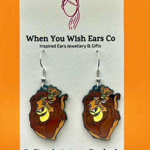 May include: A pair of dangle earrings featuring a cartoon image of Simba and Nala from the Lion King movie. The earrings are made of metal and have a silver finish. The image is colorful and detailed. The earrings are for sale on Etsy, Instagram, and Facebook.