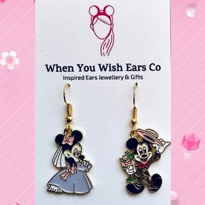 May include: A pair of gold-tone dangle earrings featuring a cartoon bride and groom Mickey Mouse. The bride Mickey Mouse is wearing a white dress and veil, while the groom Mickey Mouse is wearing a brown suit and a hat. The earrings are on a white card with the text "When You Wish Ears Co. Inspired Ears Jewellery & Gifts. On Etsy Instagram Facebook."