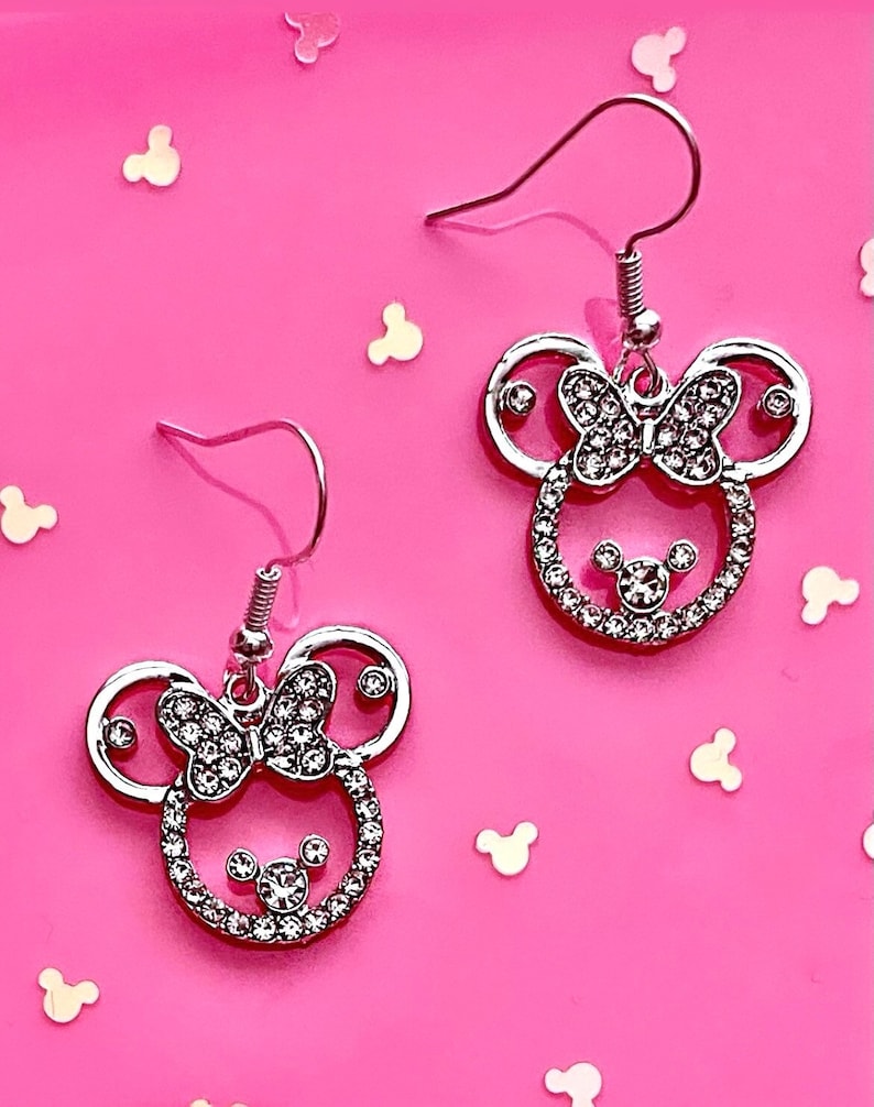 May include: A pair of silver earrings with a rhinestone-encrusted Minnie Mouse design. The earrings feature a bow and Mickey Mouse ears.