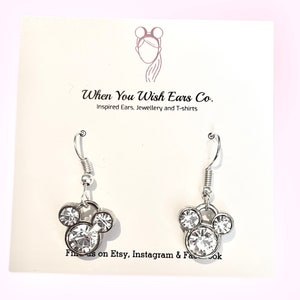 Puede incluir: Pendientes de plata con un dise&ntilde;o de cabeza de Mickey Mouse de cristal transparente. Los pendientes est&aacute;n en una tarjeta blanca con el texto "When You Wish Ears Co. Inspired Ears, Jewellery and T-shirts" y "Find us on Etsy, Instagram & Facebook".