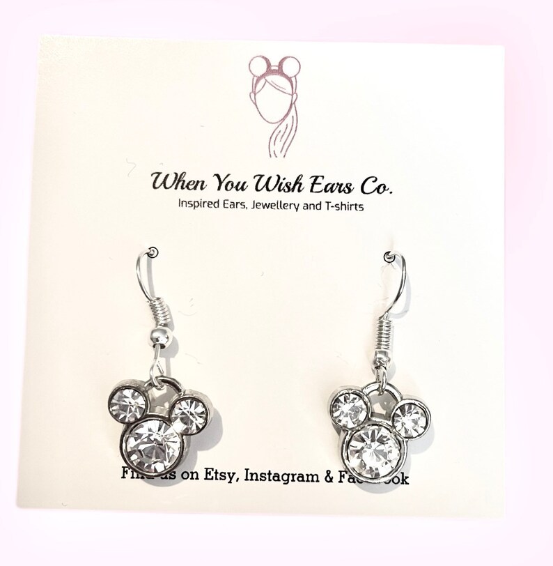 Puede incluir: Pendientes de plata con un dise&ntilde;o de cabeza de Mickey Mouse de cristal transparente. Los pendientes est&aacute;n en una tarjeta blanca con el texto "When You Wish Ears Co. Inspired Ears, Jewellery and T-shirts" y "Find us on Etsy, Instagram & Facebook".