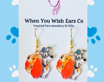 May include: Gold dangle earrings featuring Lady and the Tramp, a Disney animated film. The earrings are inspired by the characters Lady and Tramp, with Lady in red and Tramp in black and white.