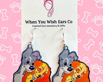 May include: A pair of dangle earrings featuring a cartoon image of Lady and the Tramp, the Disney characters, in a loving embrace. The earrings are made of silver metal and have a clear acrylic background.