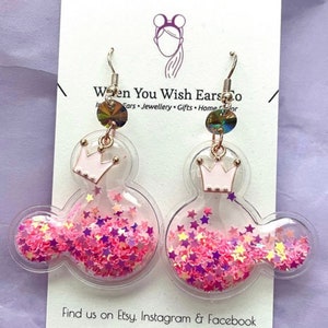 May include: A pair of pink and clear glitter filled Mickey Mouse shaped earrings with a pink crown charm. The earrings are on a white card with the text "When You Wish Ears Co. Ears Jewellery Gifts Home Decor" and "Find us on Etsy. Instagram & Facebook".