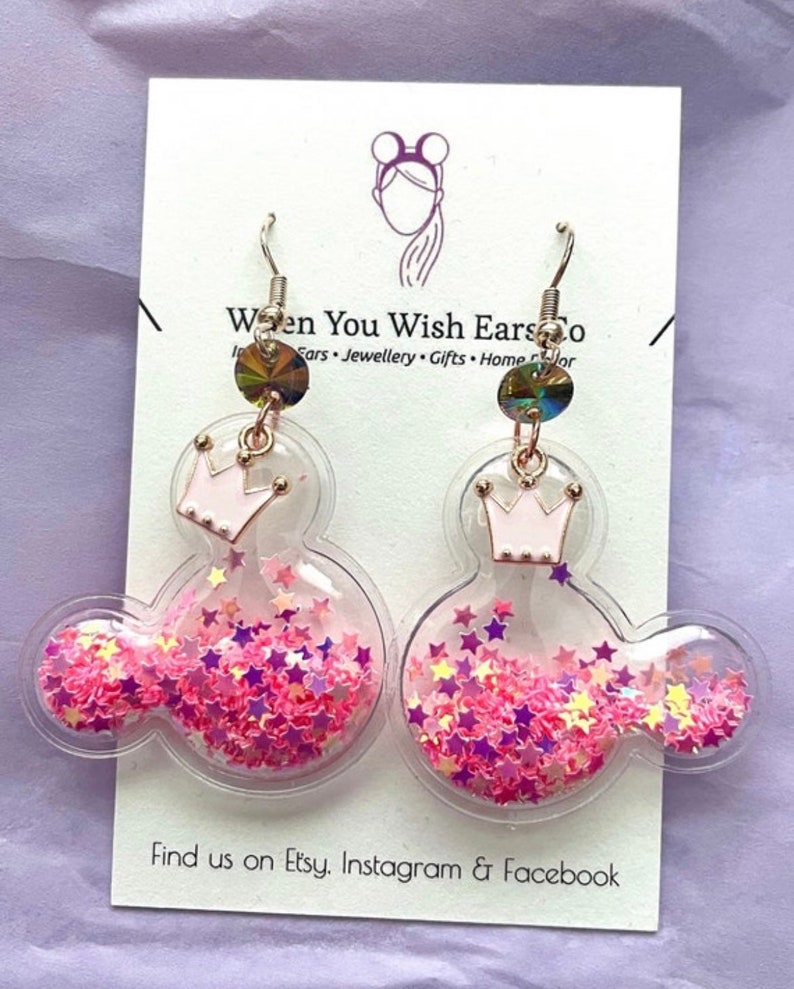 May include: A pair of pink and clear glitter filled Mickey Mouse shaped earrings with a pink crown charm. The earrings are on a white card with the text "When You Wish Ears Co. Ears Jewellery Gifts Home Decor" and "Find us on Etsy. Instagram & Facebook".