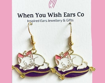 May include: Gold dangle earrings featuring a white cat with pink bow sleeping on a purple pillow. The earrings are from When You Wish Ears Co. Inspired Ears Jewellery & Gifts. On Etsy, Instagram, Facebook.