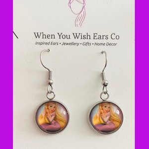 May include: A pair of silver earrings with a round glass charm featuring a cartoon image of Rapunzel from the Disney movie Tangled. The earrings are on a white card with the text "When You Wish Ears Co. Inspired Ears Jewellery Gifts Home Decor" and "Find us on Etsy, Instagram & Facebook".