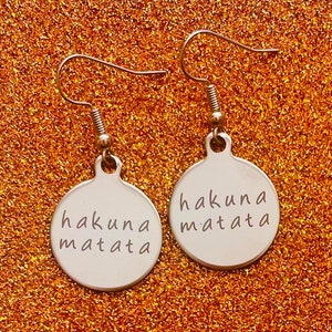Könnte beinhalten: Ein Paar silberne Ohrringe mit runder Scheibe. Jede Scheibe trägt die Aufschrift "hakuna matata".