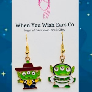 May include: A pair of gold-tone earrings featuring a green alien character from the Toy Story movie franchise. The alien is wearing a red cowboy hat and a yellow vest. The earrings are on a white background with the text "When You Wish Ears Co. Inspired Ears Jewellery & Gifts" and "On Etsy Instagram Facebook".