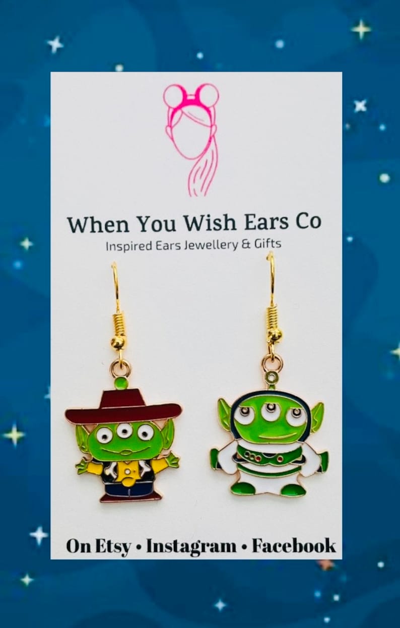 May include: A pair of gold-tone earrings featuring a green alien character from the Toy Story movie franchise. The alien is wearing a red cowboy hat and a yellow vest. The earrings are on a white background with the text "When You Wish Ears Co. Inspired Ears Jewellery & Gifts" and "On Etsy Instagram Facebook".
