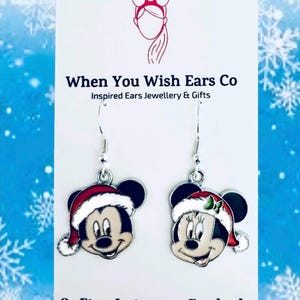May include: A pair of silver dangle earrings featuring a Christmas-themed Mickey Mouse and Minnie Mouse. Mickey is wearing a red Santa hat with white trim and Minnie is wearing a red Santa hat with white trim and a green bow. The earrings are on a white card with the text "When You Wish Ears Co Inspired Ears Jewellery & Gifts On Etsy Instagram Facebook".