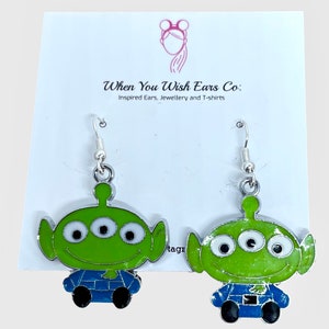 May include: A pair of silver dangle earrings featuring a green alien character with three eyes and a blue jumpsuit. The character is from the Toy Story movie franchise.