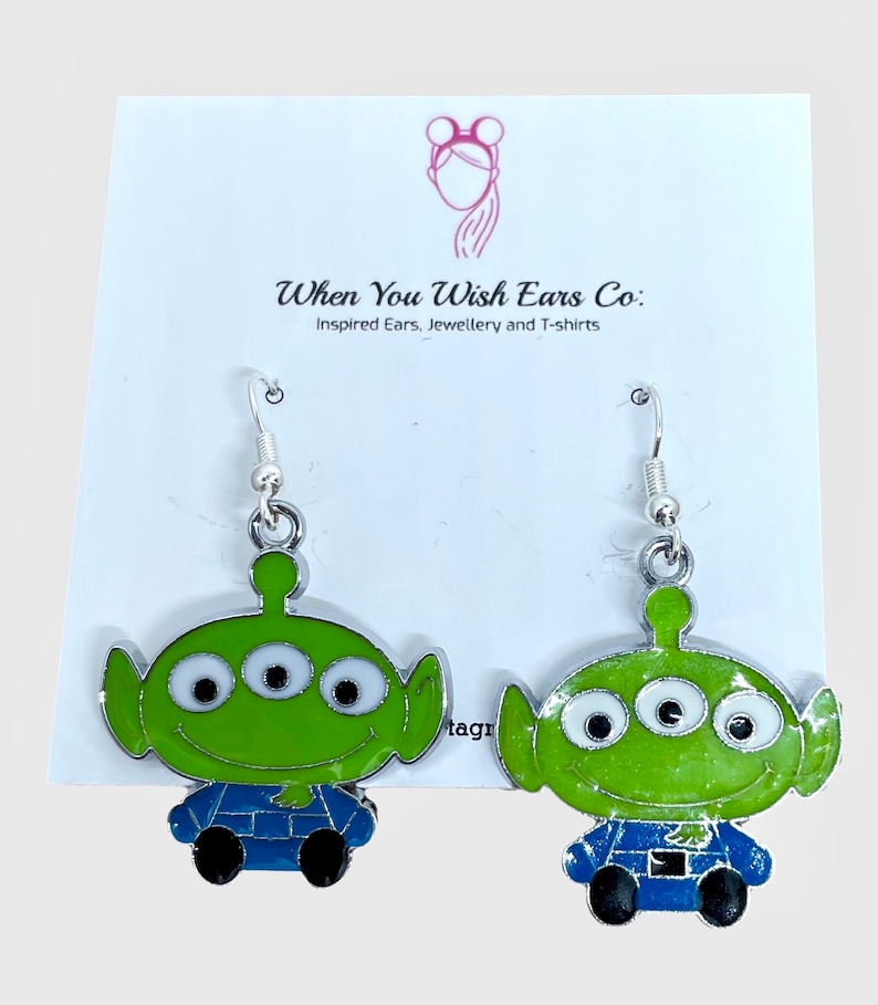 May include: A pair of silver dangle earrings featuring a green alien character with three eyes and a blue jumpsuit. The character is from the Toy Story movie franchise.